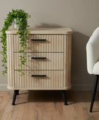 Wooden side table with three drawers next to a white armchair on a wooden floor.