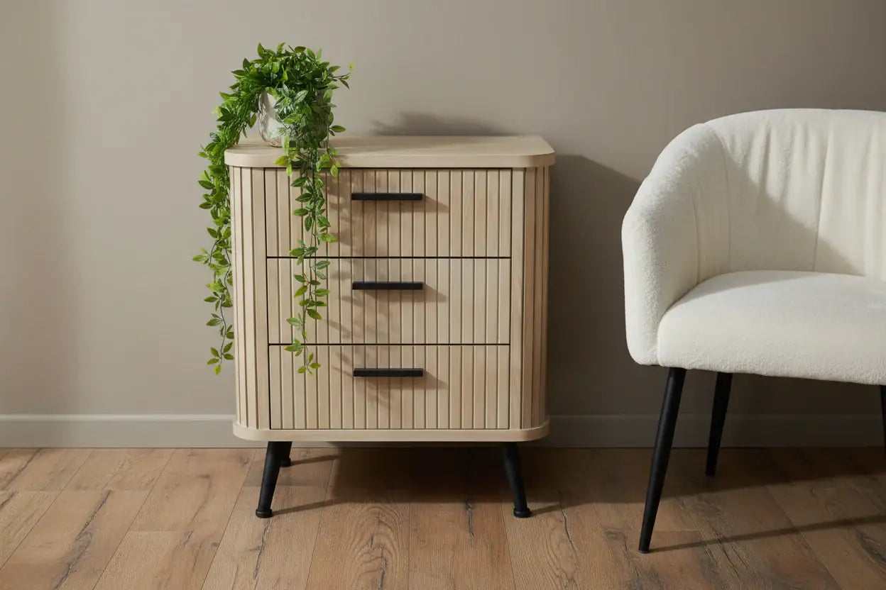 Wooden side table with three drawers next to a white armchair on a wooden floor.