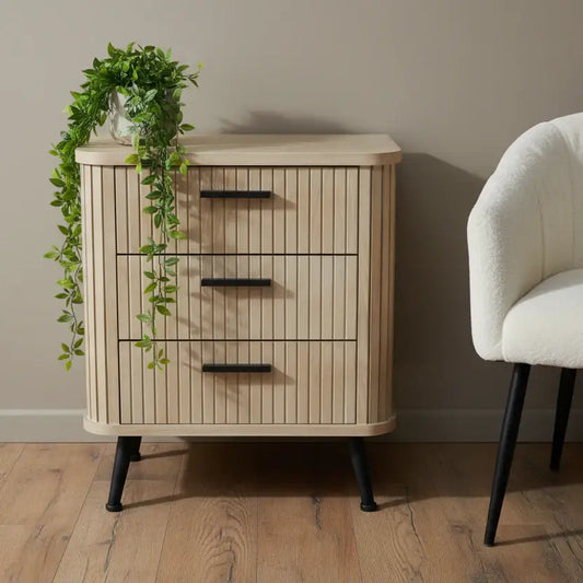 Wooden cabinet with black legs and greenery on a wooden floor, with a chair in the background.
