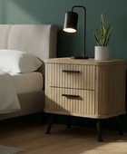 Wooden nightstand with lamp and plant next to a bed in a bedroom.