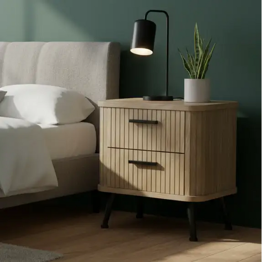 Wooden nightstand with lamp and plant next to a bed in a bedroom.