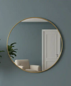 Round gold-framed mirror on a dark teal wall with a white door and plant in the reflection.