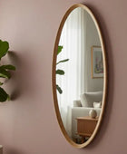 Oval mirror with wooden frame on a pink wall with a plant and part of a room visible in the reflection.
