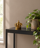 A small ornate gold crown figurine sits on a black console table next to a potted green plant against a warm beige wall.