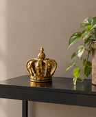 A decorative gold crown sculpture sits on a black side table next to a green potted plant against a beige wall.
