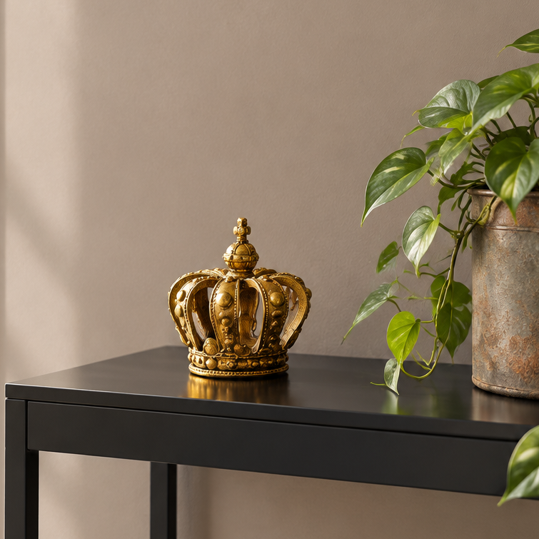 A decorative gold crown sculpture sits on a black side table next to a green potted plant against a beige wall.