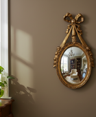 Decorative gold mirror with a bow on a wall, reflecting a room with furniture.