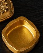 A square gold-toned metal container or dish with rounded corners and a textured metallic finish, shown on a light neutral surface with part of another gold object visible in the upper left.