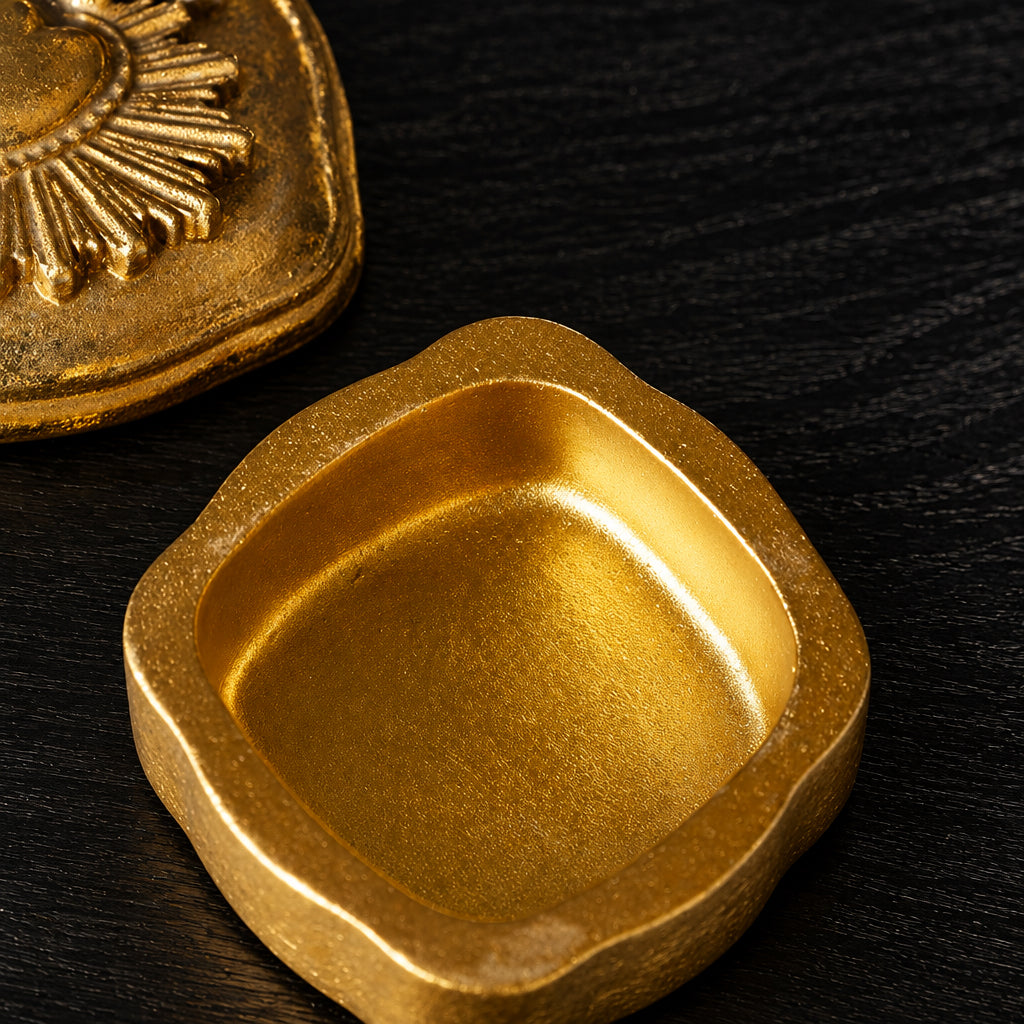 A square gold-toned metal container or dish with rounded corners and a textured metallic finish, shown on a light neutral surface with part of another gold object visible in the upper left.