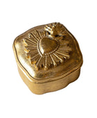 A small decorative gold-toned metal box with a hinged lid, featuring an embossed Sacred Heart design with a leaf on top, set against a light marble background.