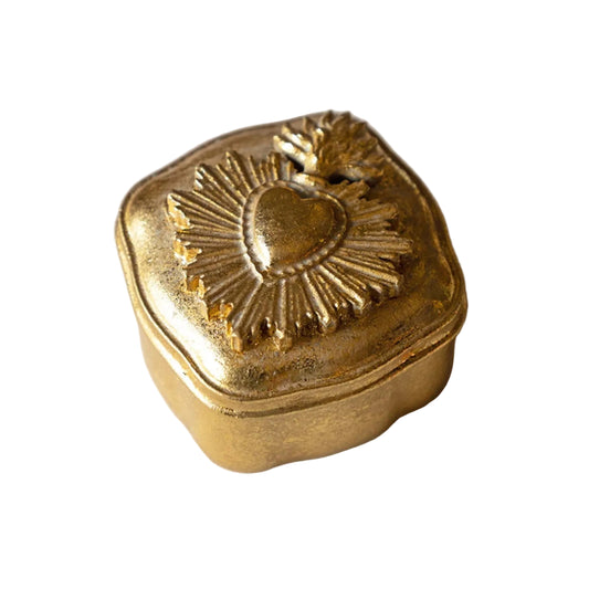A small decorative gold-toned metal box with a hinged lid, featuring an embossed Sacred Heart design with a leaf on top, set against a light marble background.