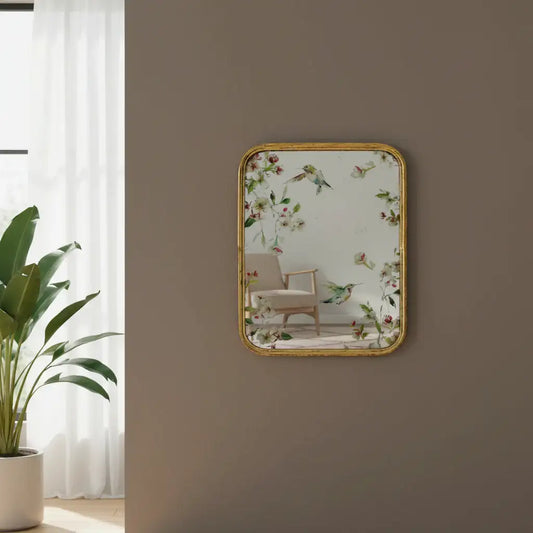 Decorative mirror with floral and bird design on a wall, with a plant and curtain in the background.