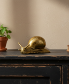 Gold snail ornament on a wooden surface with a potted plant and book in the background.
