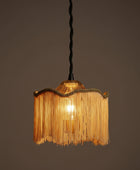 A hanging pendant light with a fringed shade made of warm gold-toned raffia or straw-like strips, a scalloped braided trim, and a frosted white socket at the top against a plain white background.