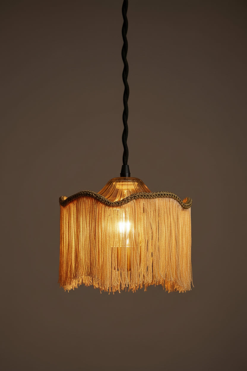 A hanging pendant light with a fringed shade made of warm gold-toned raffia or straw-like strips, a scalloped braided trim, and a frosted white socket at the top against a plain white background.