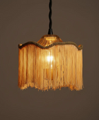 A hanging pendant light with a warm glowing bulb inside a fringed shade. The shade is made of golden-yellow fringe with a braided trim, suspended from a black cord against a plain dark brown background.