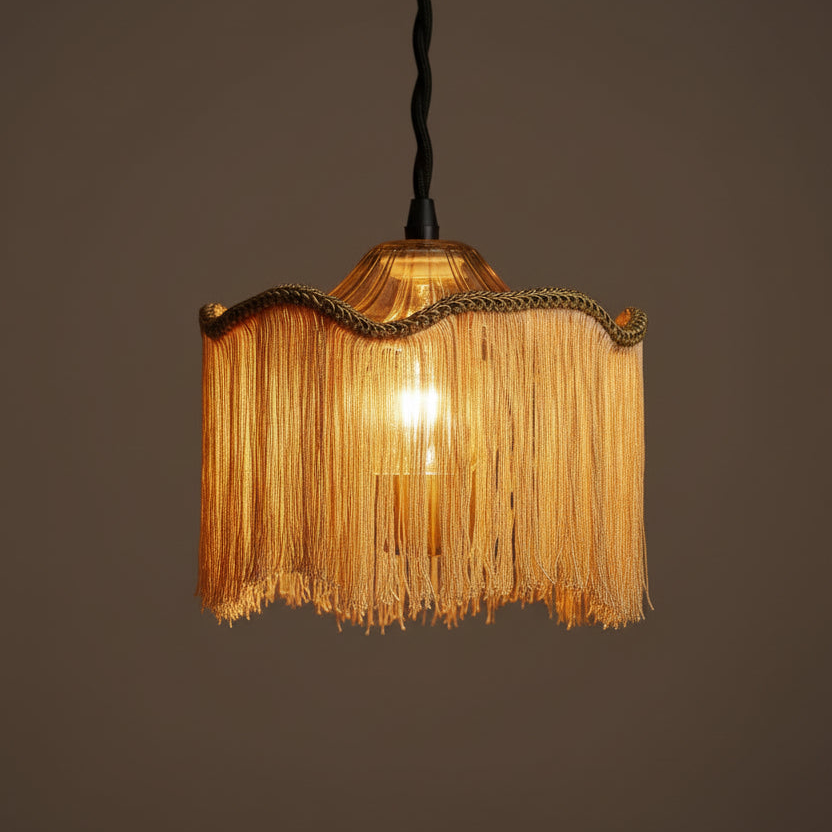 A hanging pendant light with a warm glowing bulb inside a fringed shade. The shade is made of golden-yellow fringe with a braided trim, suspended from a black cord against a plain dark brown background.