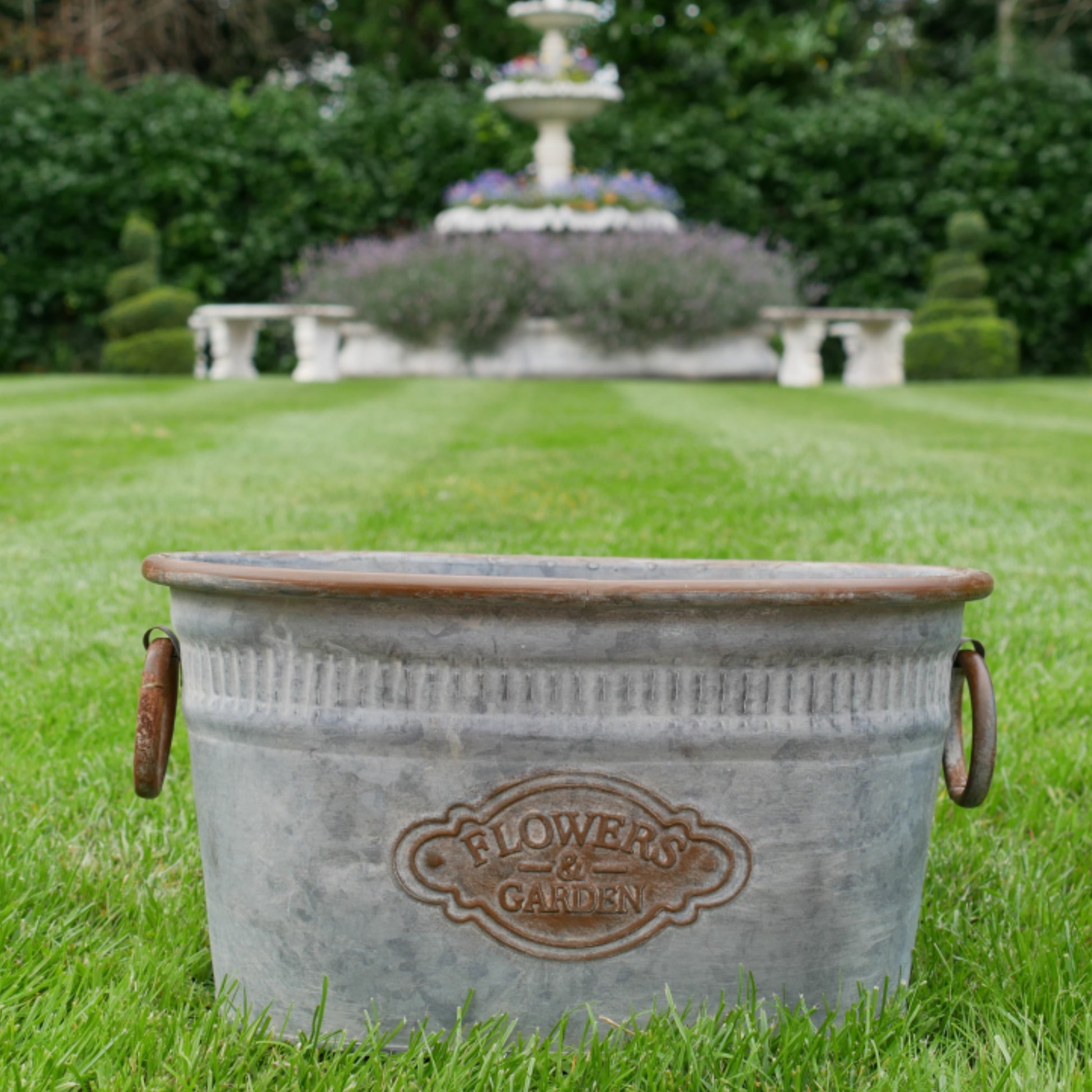 Metal flower pot with 'Flowers & Garden' branding on a grassy lawn with a fountain in the background.