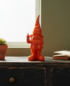 Orange gnome figurine on a wooden surface with a plant and books in the background.