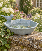 A light blue-gray hexagonal garden birdbath with a small bird perched on the left side, filled with water and set on a stone wall in a lush garden with flowers and ivy.
