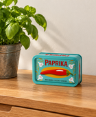 A turquoise metal tin labeled 'PAPRIKA' sits on a wooden table beside a potted basil plant against a plain wall.