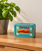 A decorative tin canister labeled 'PAPRIKA' sits on a wooden table next to a potted green basil plant against a plain wall.