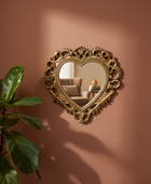An ornate gold heart-shaped wall mirror with a decorative carved frame, mounted on a warm terracotta-colored wall. A green potted plant is visible on the left, and the mirror reflects a softly lit living room interior.