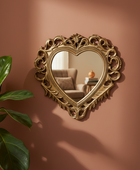 An ornate heart-shaped decorative wall mirror with an intricate gold-toned scrollwork frame, mounted on a warm terracotta-colored wall. A green houseplant is visible in the lower-left corner, and the mirror reflects a softly lit living room interior.