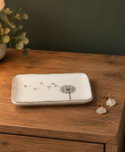 A rectangular white ceramic jewelry tray with a black dandelion design, styled on a wooden nightstand next to a pair of gold and white earrings.