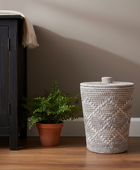 Woven laundry basket next to a plant and dark wooden cabinet against a beige wall.