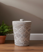 Woven laundry basket with a lid on a wooden floor next to a potted plant against a neutral wall.