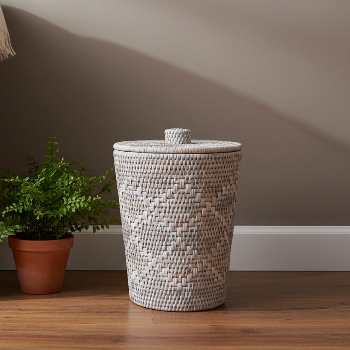 Woven laundry basket with a lid on a wooden floor next to a potted plant against a neutral wall.