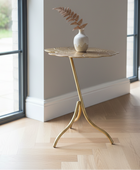 A gold metal leaf-shaped side table with a small white vase containing a dried fern, set on a light wood herringbone floor in a bright room with large windows.