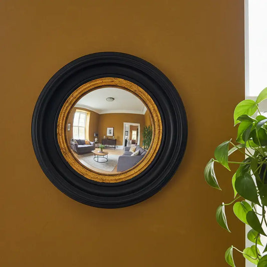 Round mirror with black frame on a mustard yellow wall, reflecting a living room.