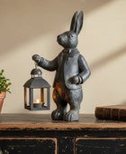 Decorative rabbit figurine holding a lantern on a wooden surface with a plant and book in the background.