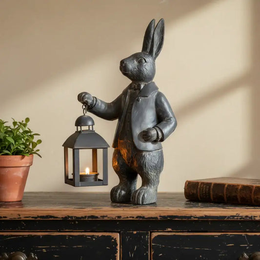 Decorative rabbit figurine holding a lantern on a wooden surface with a plant and book in the background.