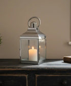 Silver lantern with a candle on a wooden surface next to a potted plant and a book.