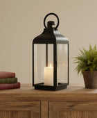 Black lantern with a candle on a wooden table next to books and a potted plant.