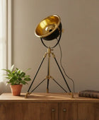 Vintage-style tripod lamp with gold bowl on a wooden surface next to a plant and book.