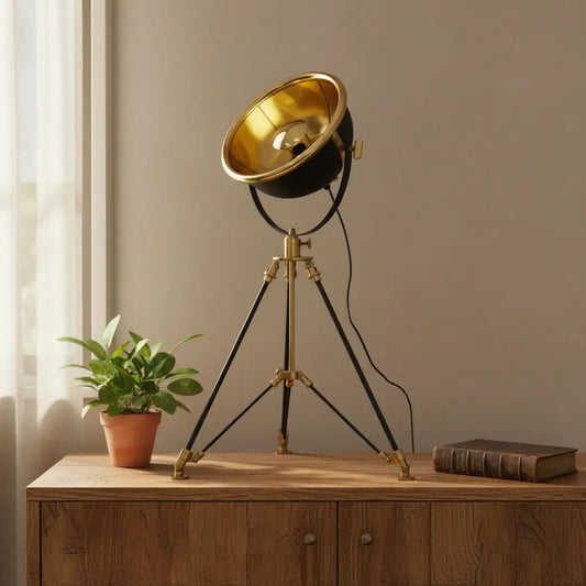 Vintage-style tripod lamp with gold bowl on a wooden surface next to a plant and book.