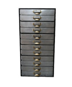 Tall metal drawer unit with brass handles against a brick wall.