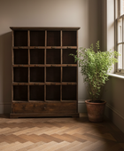 A tall dark wood cabinet with multiple open cubbies and a small drawer at the bottom, placed in a room with a herringbone wood floor, beige walls, and a potted plant beside a window.