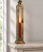 A tall, narrow brass lantern-style candle holder with a lit red pillar candle inside, displayed on a distressed white wooden cabinet against a neutral gray wall.