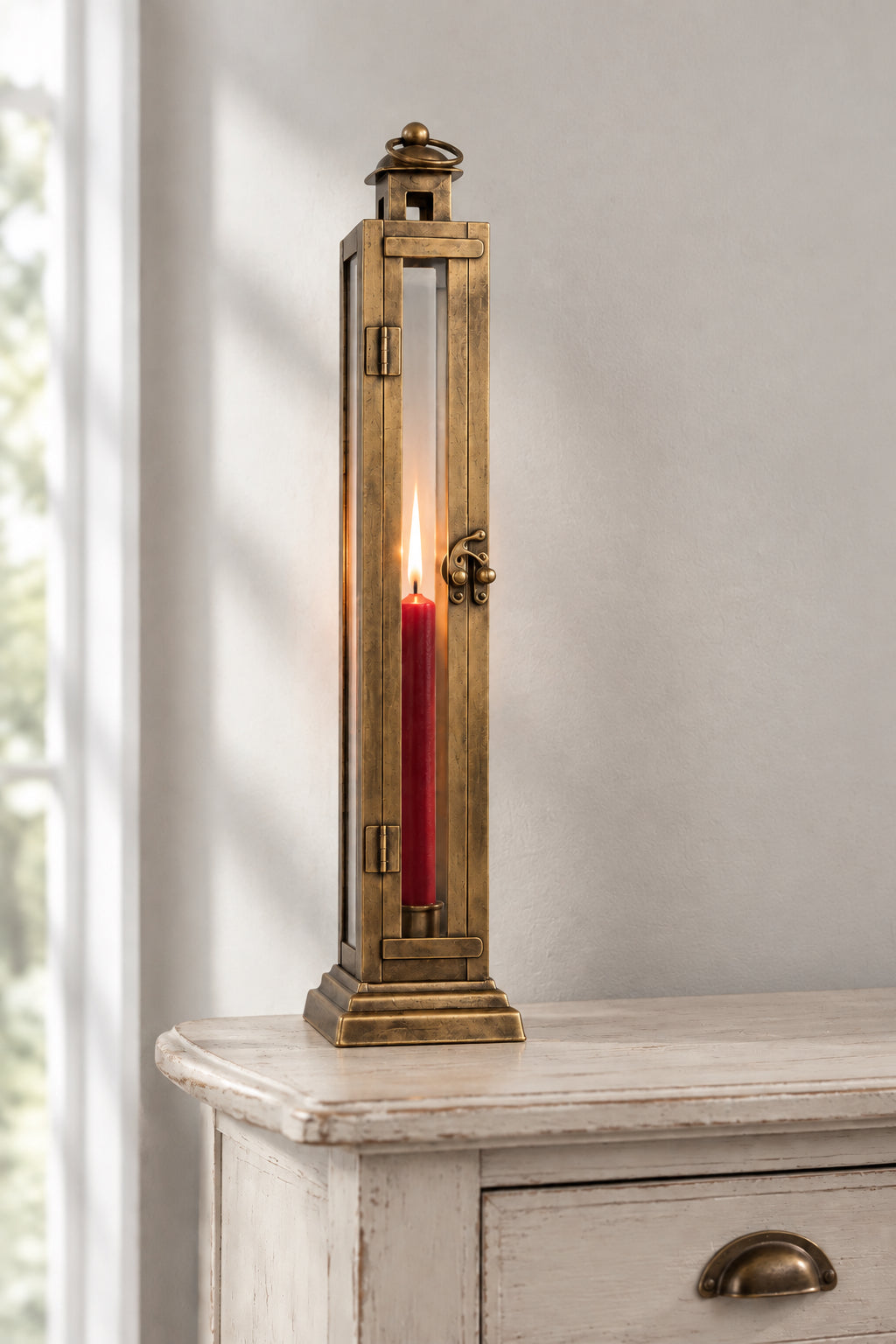 A tall, narrow brass lantern-style candle holder with a lit red pillar candle inside, displayed on a distressed white wooden cabinet against a neutral gray wall.