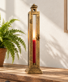 A tall rectangular brass lantern-style candle holder with glass panels, shown lit with a red taper candle inside, set against a neutral gray background.