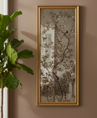 A tall, narrow framed wall art piece with a botanical print of branches, leaves, and birds, displayed on a warm taupe wall next to a green potted plant.