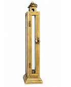 A tall, narrow brass lantern-style candle holder with a lit red pillar candle inside, displayed on a distressed white wooden cabinet against a neutral gray wall.