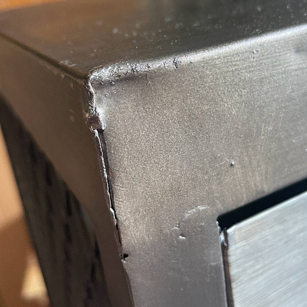 Close-up of a dark gray or black metal corner with visible scratches, dust, and a brushed metallic surface on the adjacent panel.