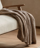Brown knitted blanket draped over a beige armchair on a wooden floor.