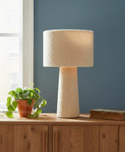 Table lamp with textured beige shade on a wooden surface next to a plant and book against a blue wall.